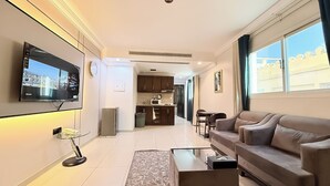 A modern living room with a sofa, coffee table, and TV. There is a kitchen area with cabinets and appliances in the background.