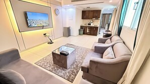 A modern living room with a flat-screen TV, a sofa set, a coffee table, and a kitchen area in the background.