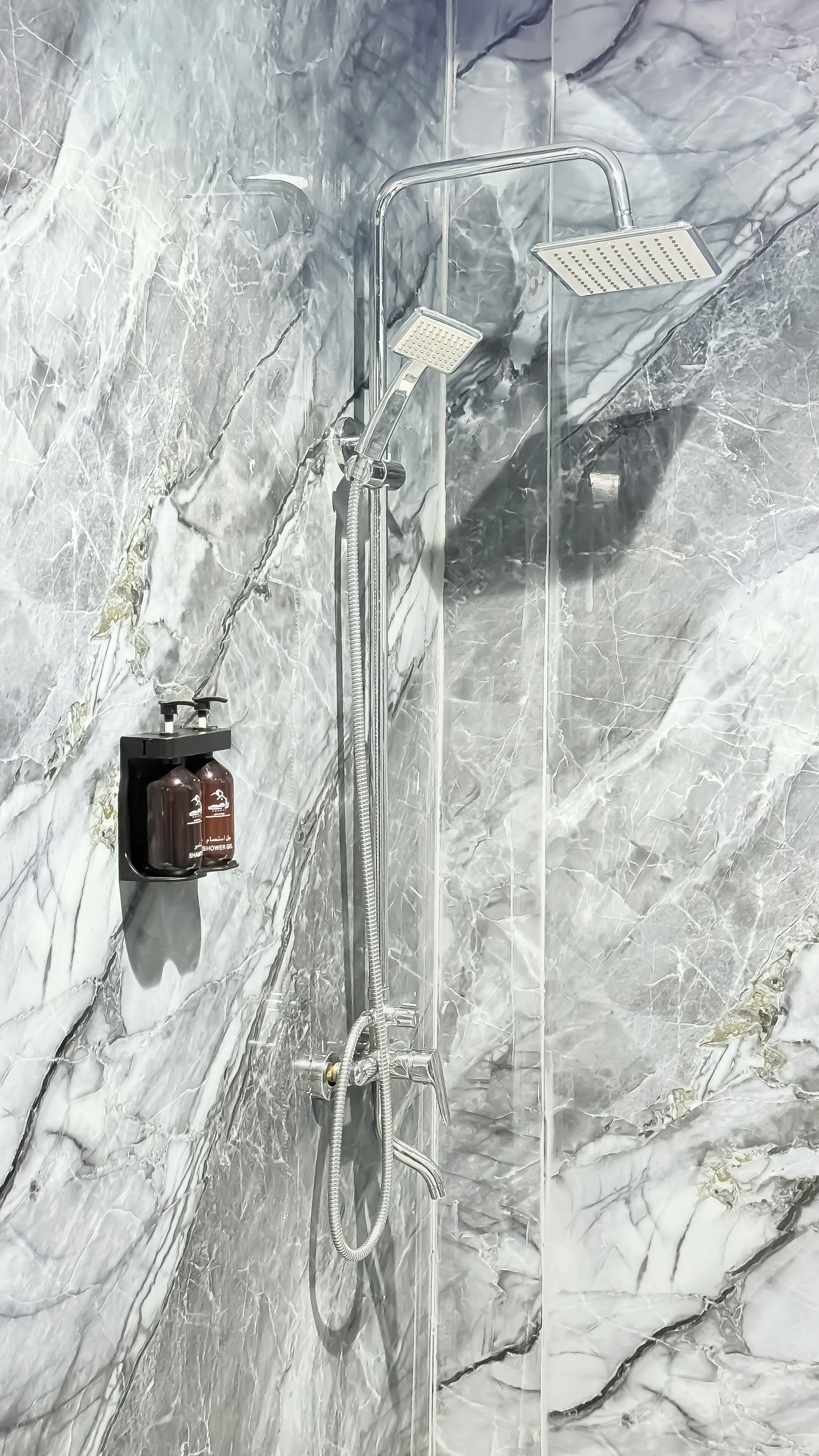 A shower with a handheld showerhead and a wall-mounted one, a control panel, and a bottle of shower gel.
