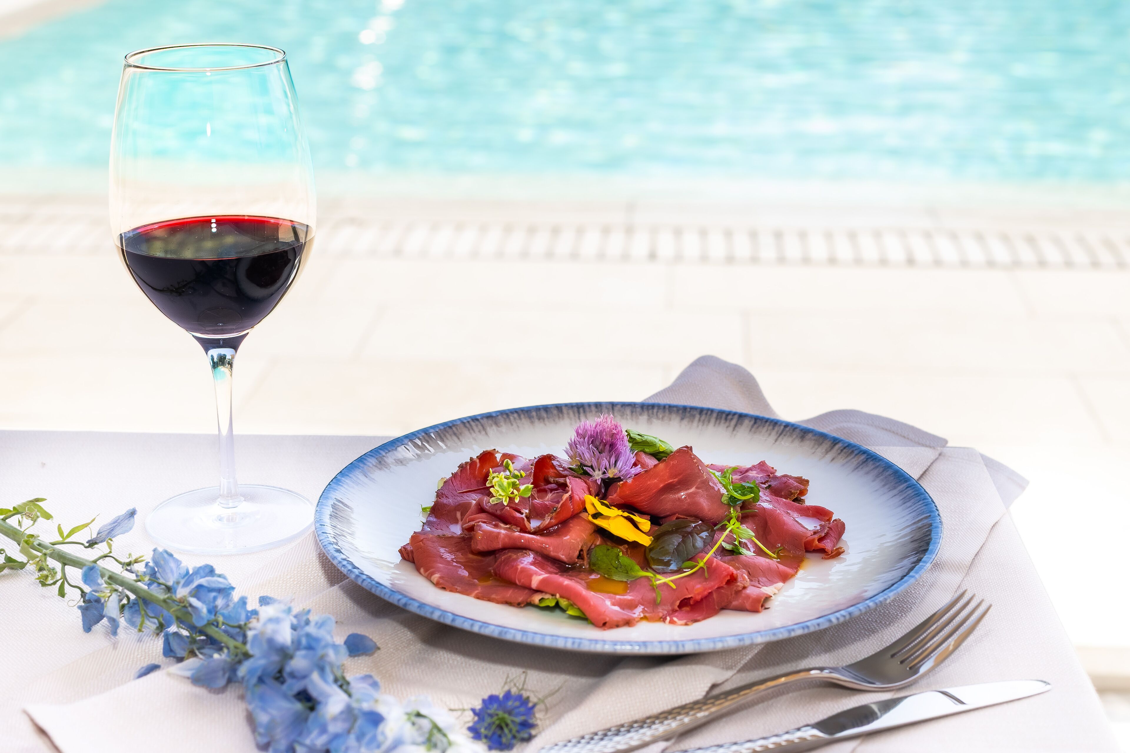 A dining table with a plate of food, a glass of red wine, and a pool in the background.