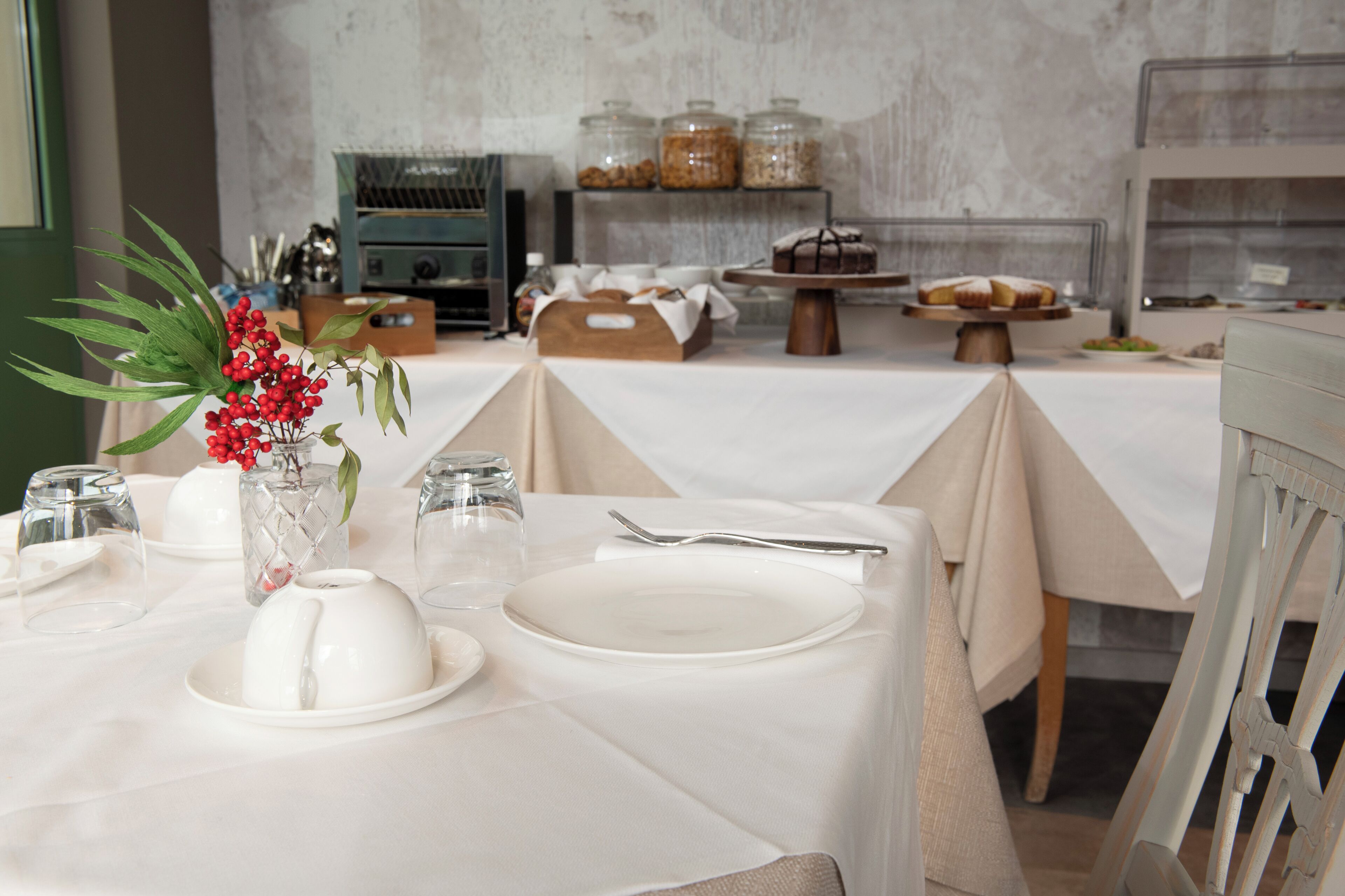 A dining area with tables set for service, a vase with red berries, and a display of baked goods in the background.