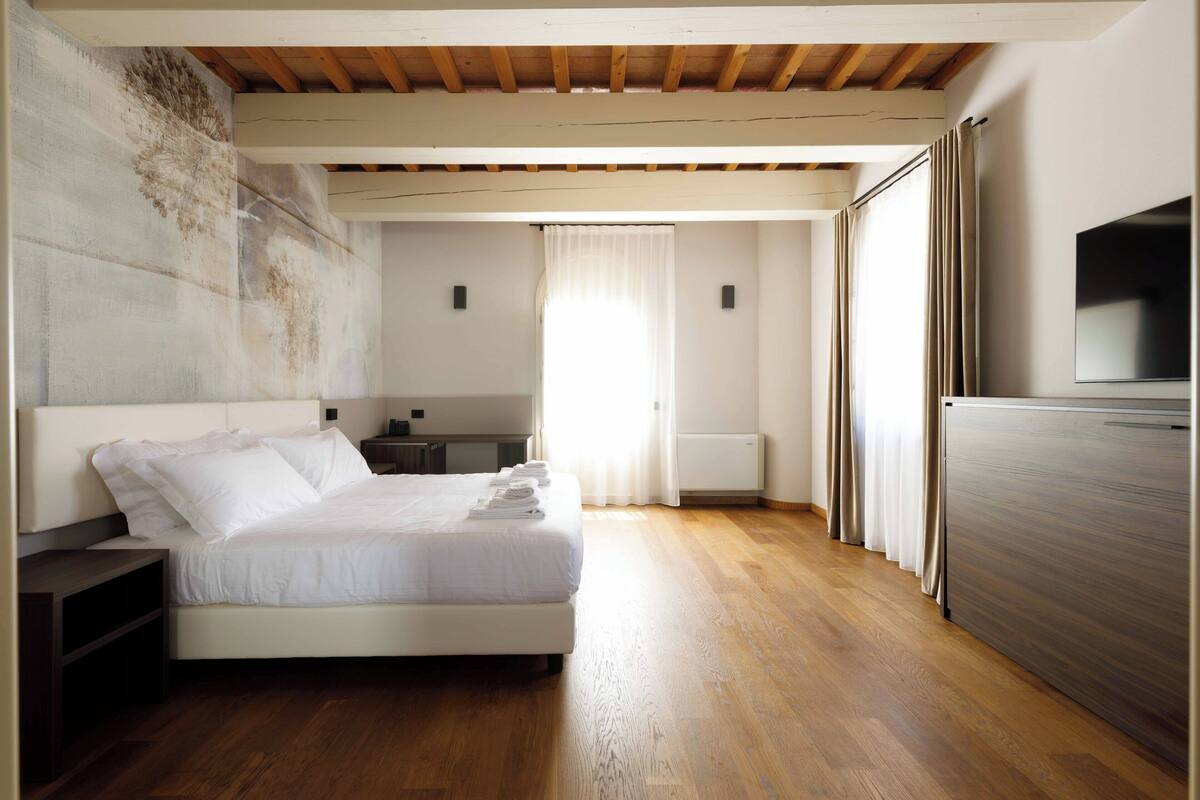 A bedroom with a bed, wooden flooring, a TV, and a window with curtains.
