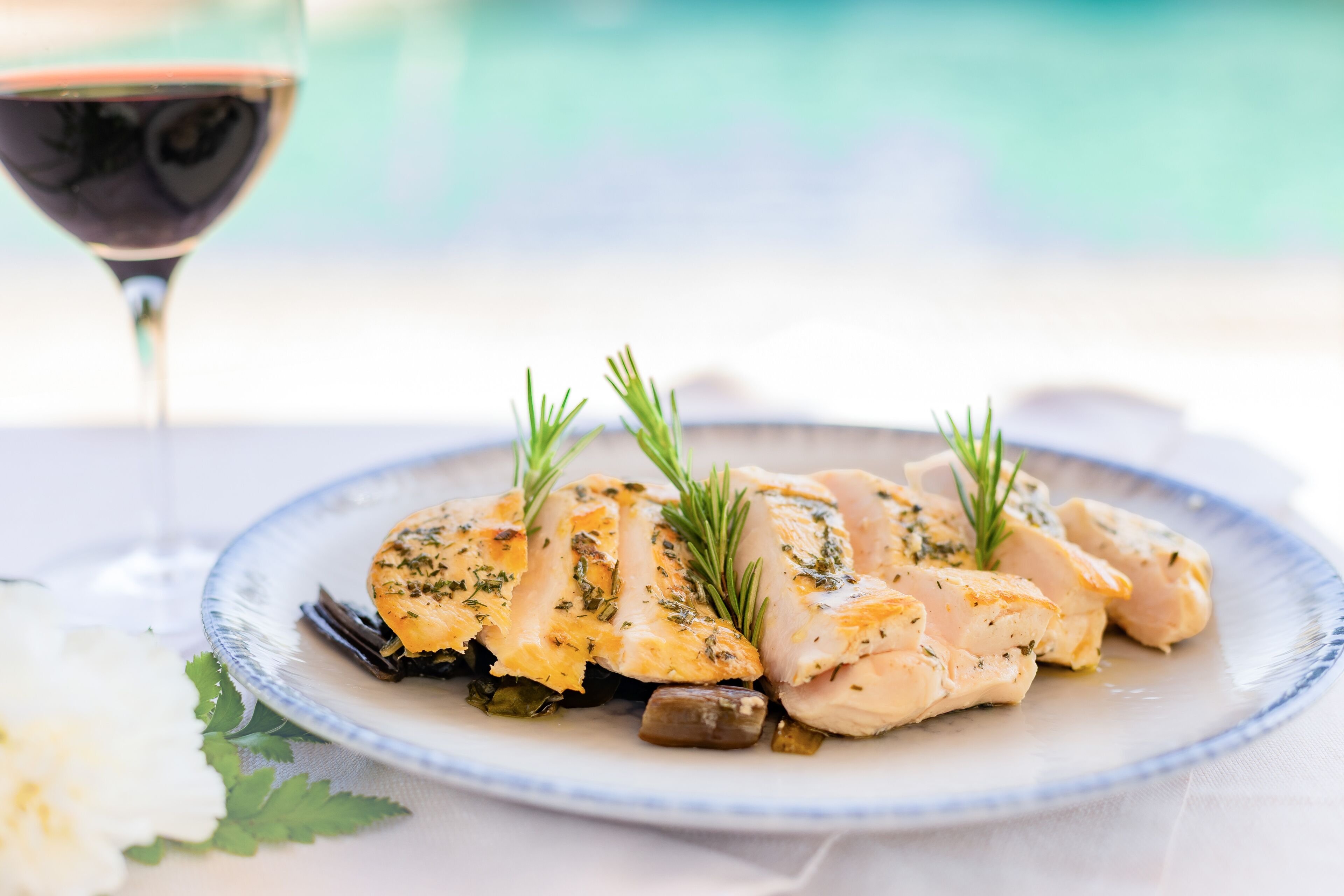 A plate of chicken with herbs and olives, a glass of red wine, and a white flower on a table.