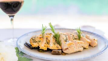 A plate of chicken with herbs and olives, a glass of red wine, and a white flower on a table.
