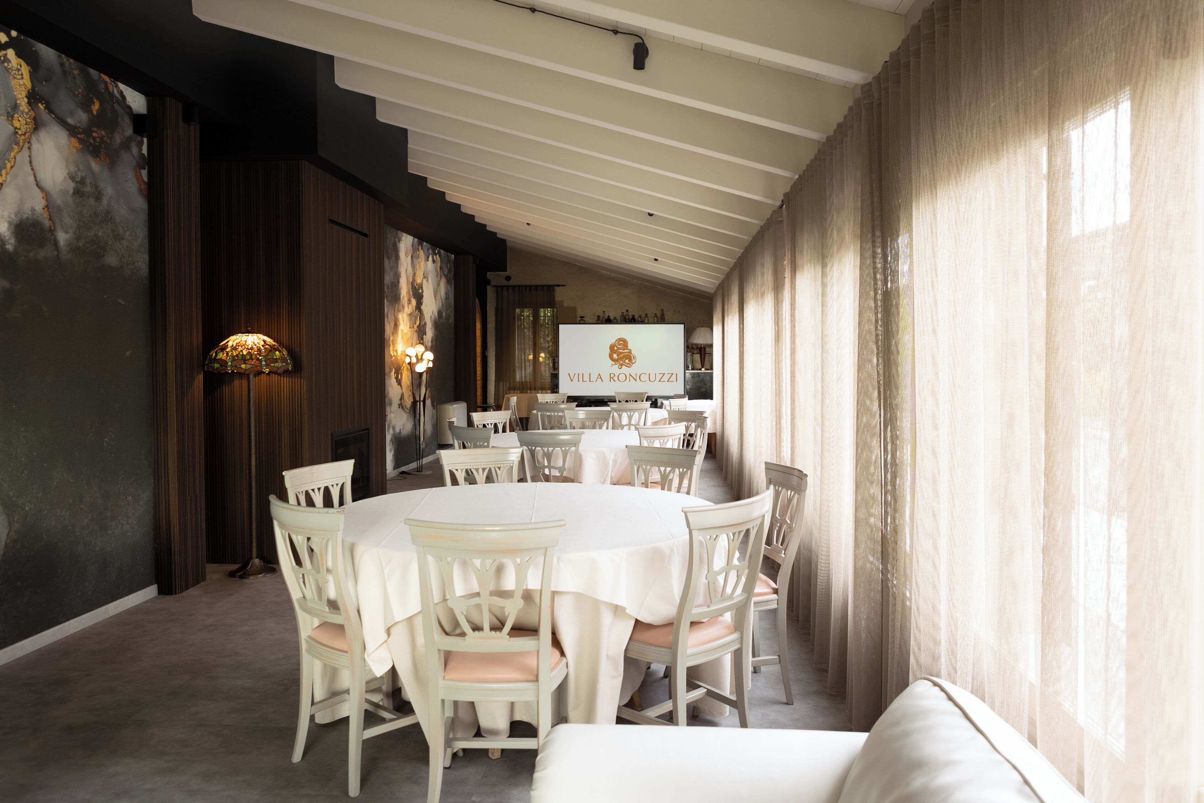 A dining area with round tables and chairs, a television screen displaying 'VILLA RONCUZZI,' and a large window with curtains.