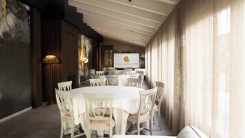A dining area with round tables and chairs, a television screen displaying 'VILLA RONCUZZI,' and a large window with curtains.