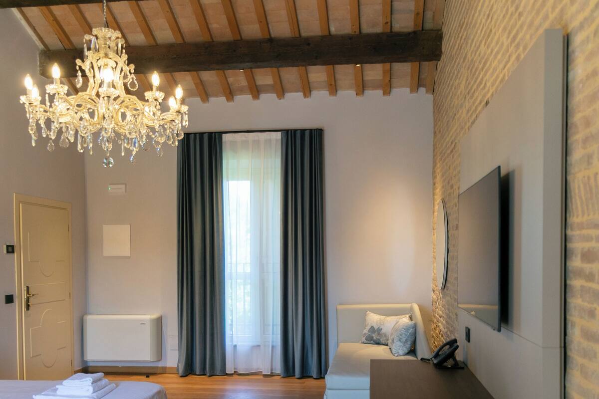 A modern hotel room with a wooden ceiling, a chandelier, a bed, a seating area, and a TV mounted on the wall.