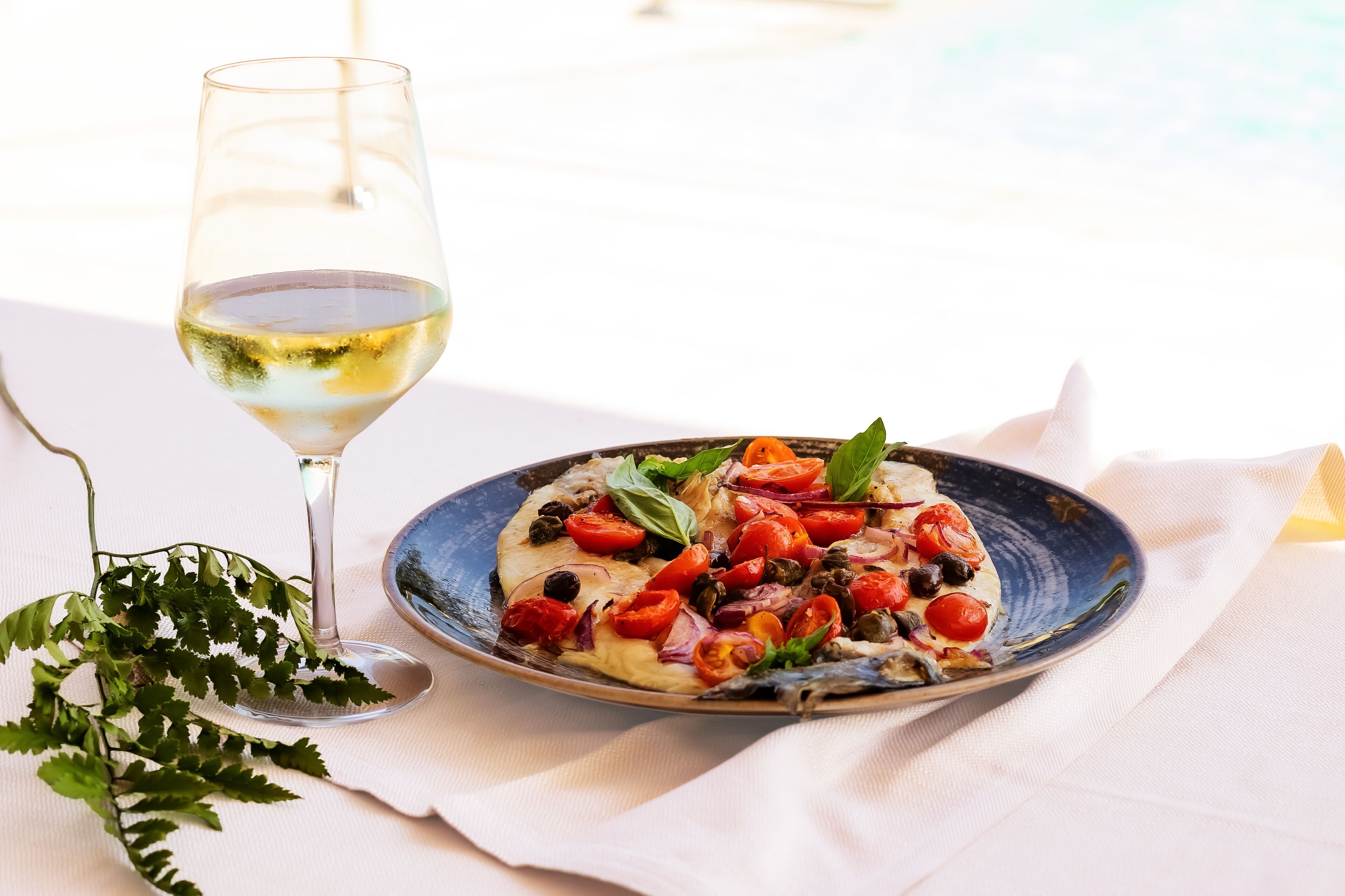 Un plato de pizza con diferentes ingredientes y una copa de vino blanco sobre una mesa.