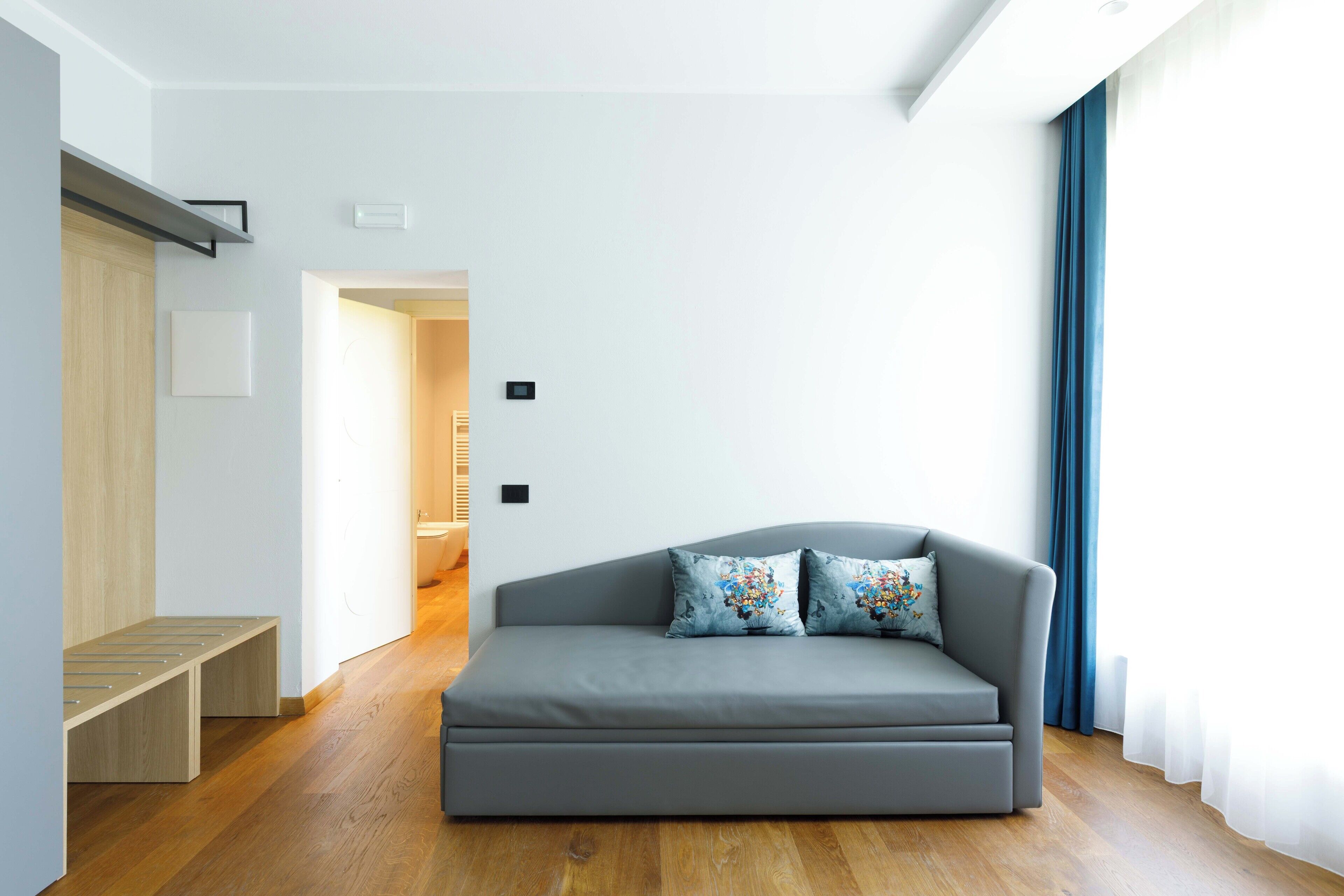 Een moderne woonkamer met een grijze bank, twee decoratieve kussens, een houten bankje en uitzicht op een badkamer door een open deur.