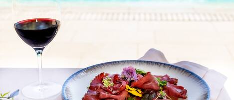 A plate of food with a glass of red wine, a pool in the background, and a napkin on the table.