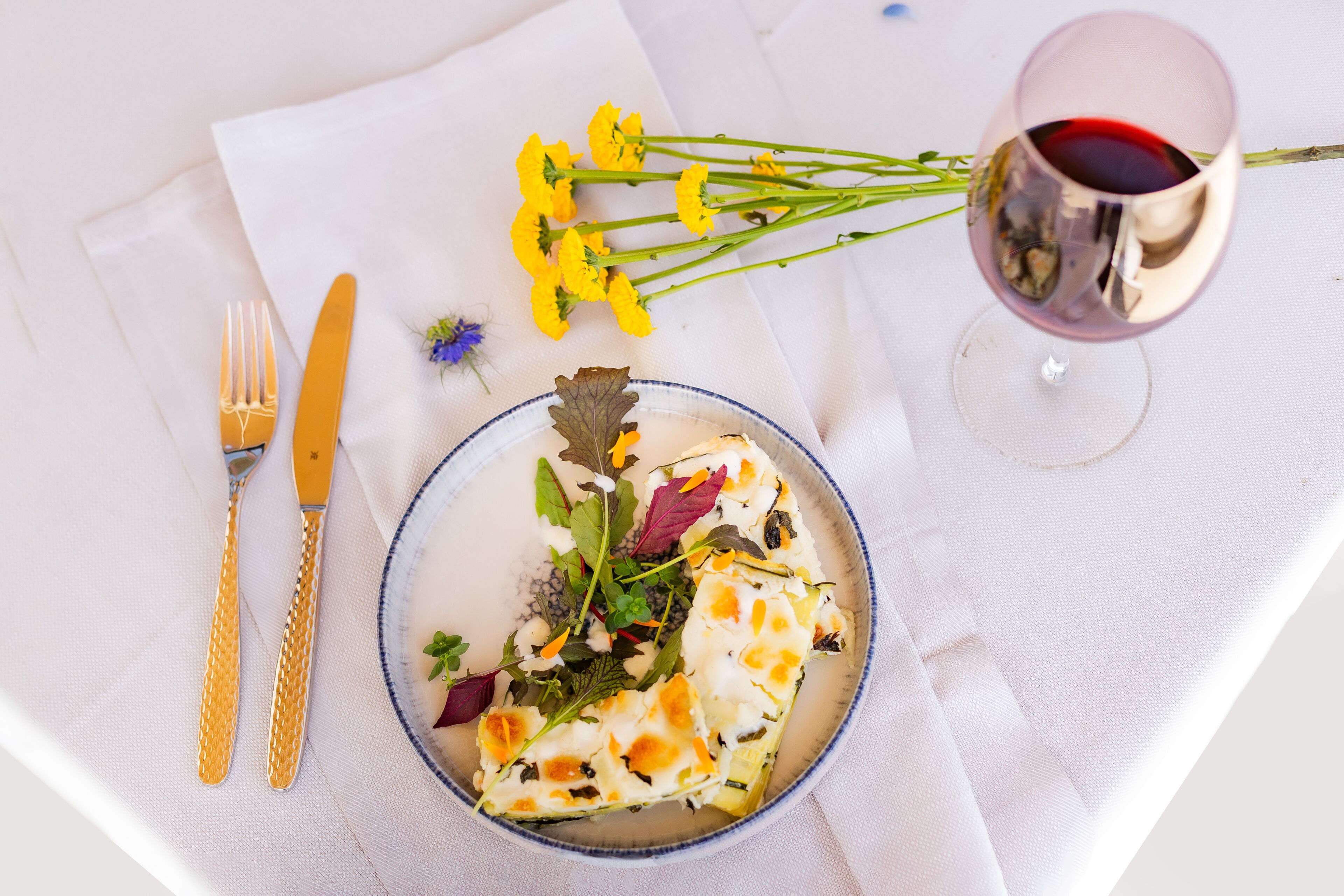 Une table blanche avec une assiette de nourriture, un verre de vin rouge et un bouquet de fleurs jaunes.