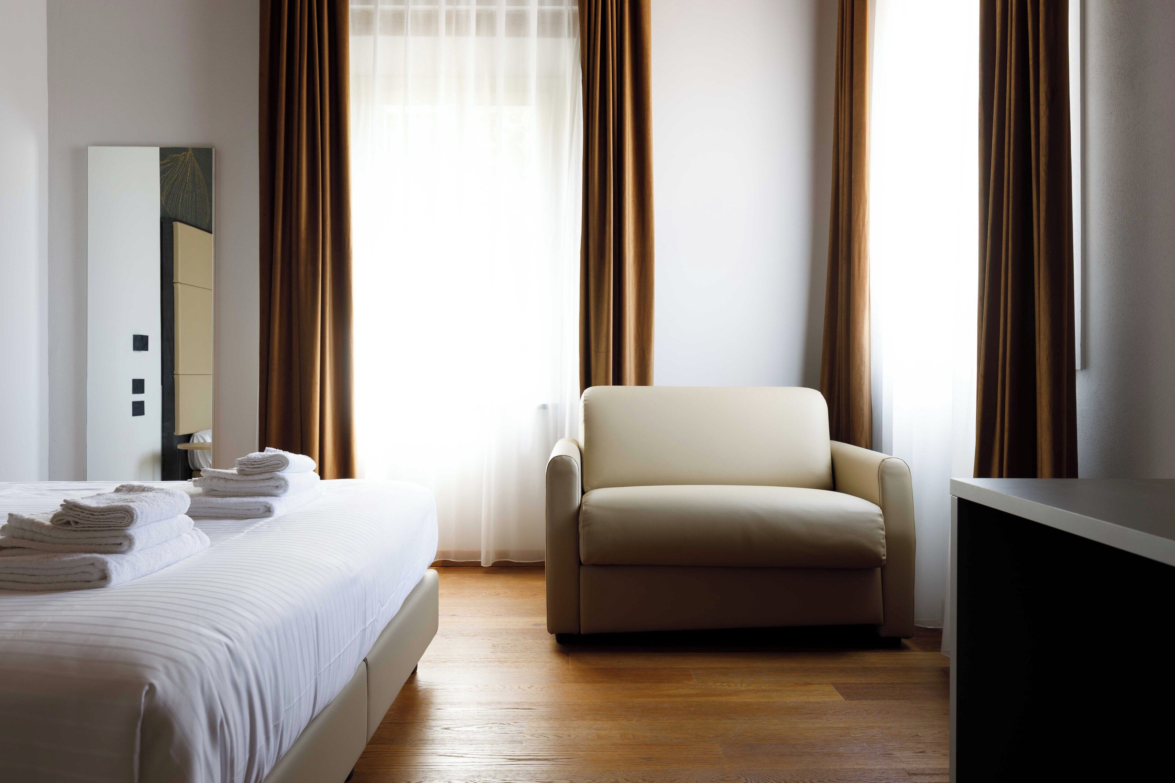 Quarto de hotel com uma cama, uma cadeira, uma escrivaninha e cortinas.