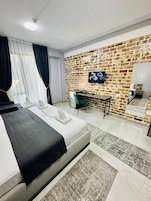 A modern hotel room with a brick wall, a bed with a black headboard, a desk with a chair, a flat-screen TV, and a large mirror.
