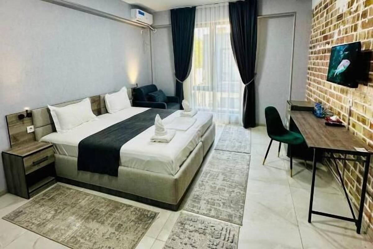 A modern hotel room with a large bed, a desk, a TV mounted on the brick wall, and a window with curtains.