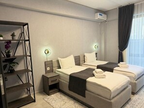 Une chambre d’hôtel moderne avec deux lits, un petit bureau, une étagère avec des plantes et une fenêtre avec des rideaux.