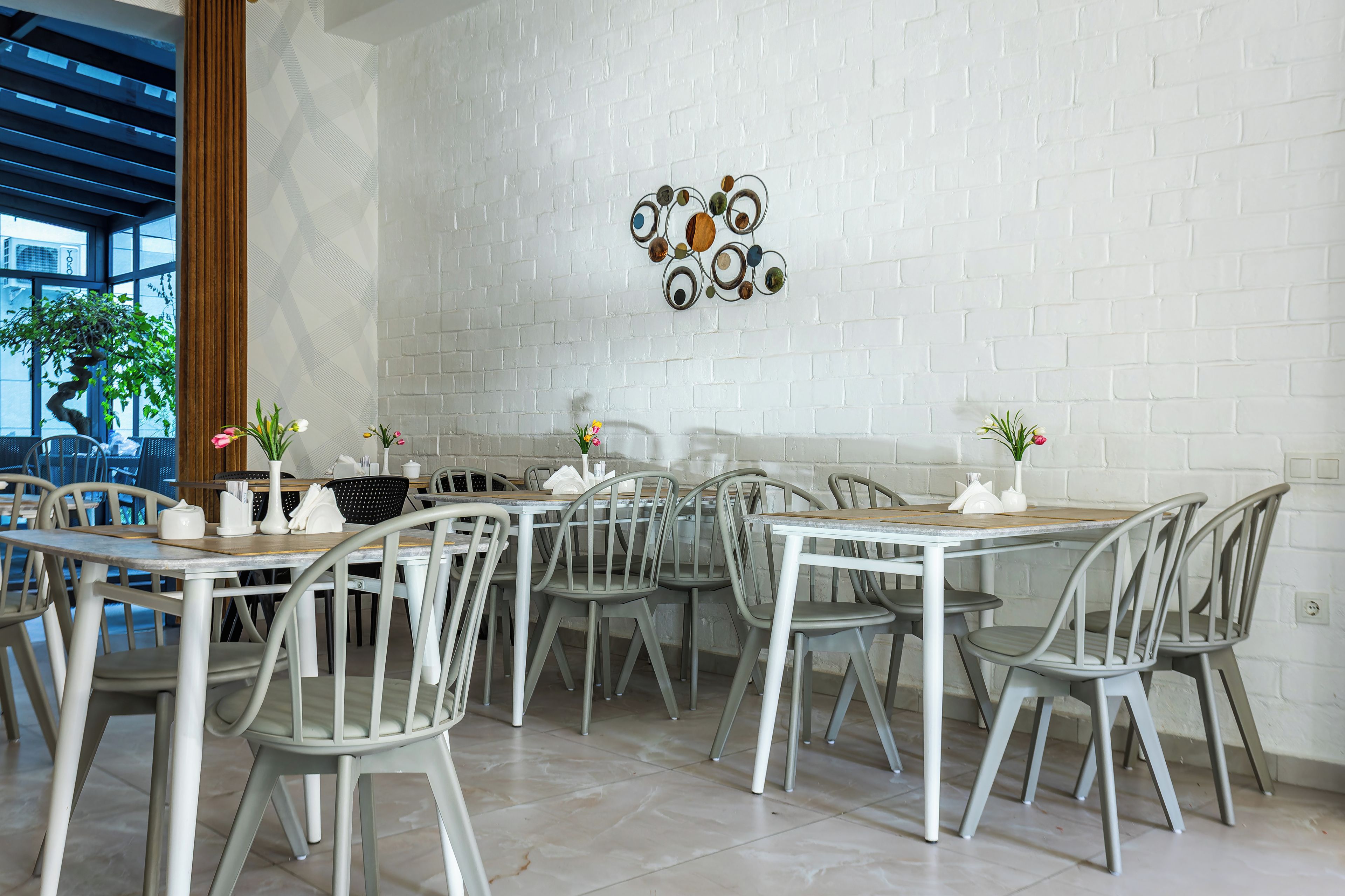 A dining area with white brick walls, grey chairs, and tables. There are decorative wall art pieces and potted plants on the tables.