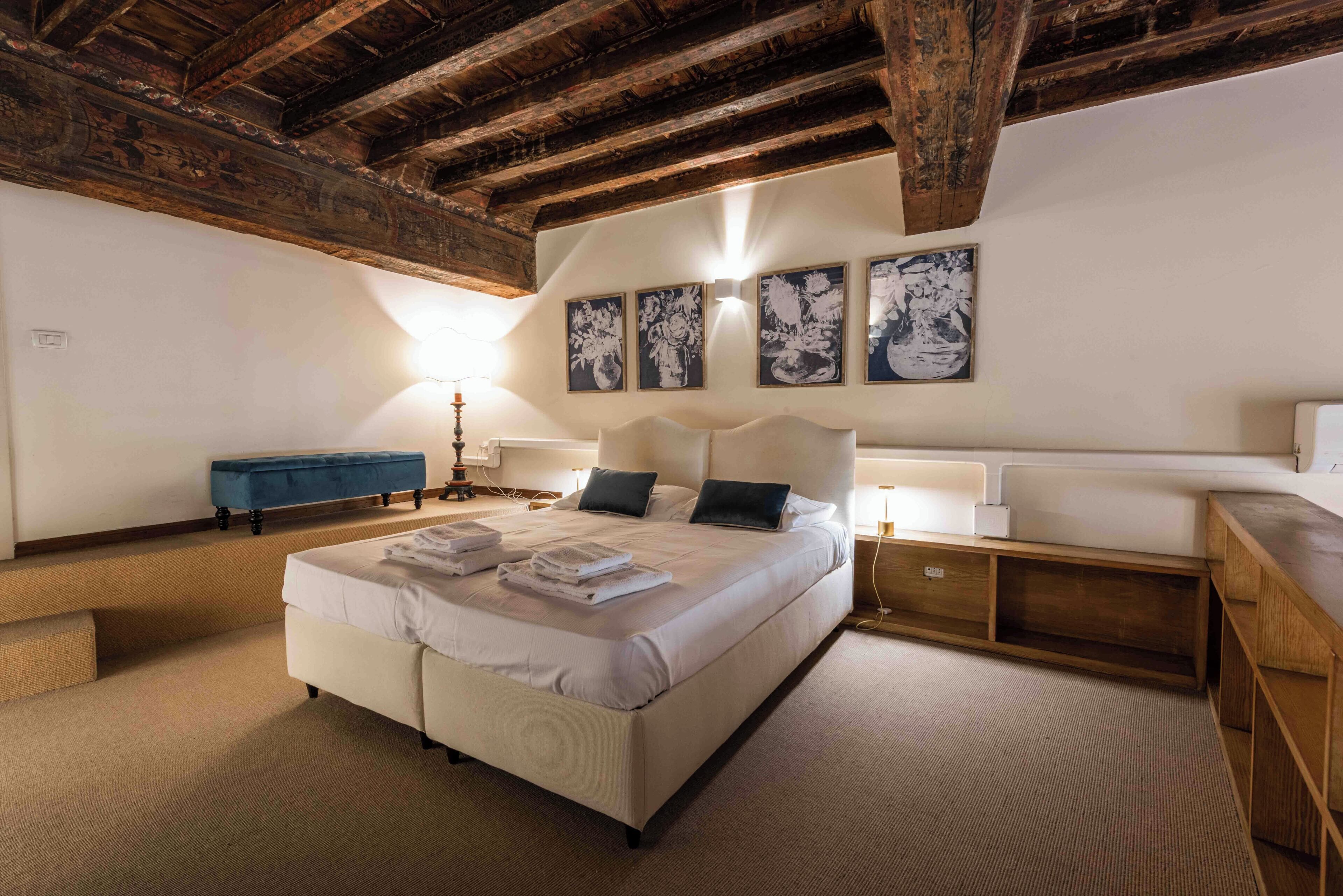 Et soveværelse med en stor seng, træbjælker, en blå bænk og kunst på væggen.