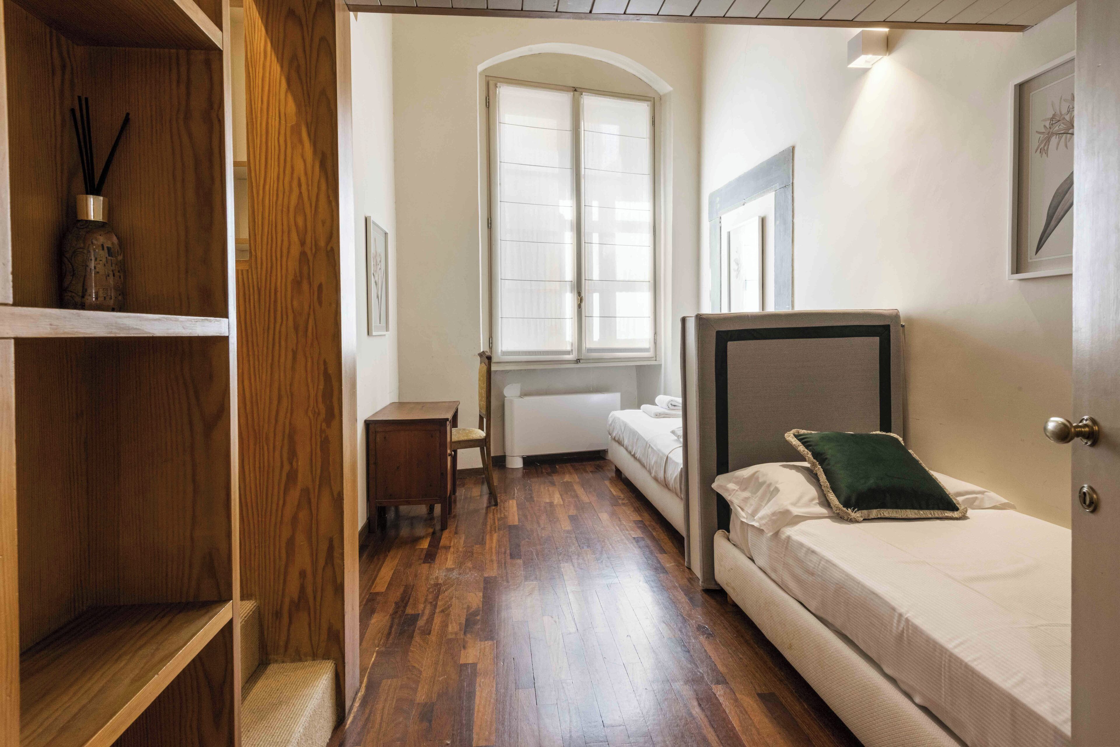 A bedroom with a wooden floor, a bed with a green pillow, a bedside table, and a window with blinds.