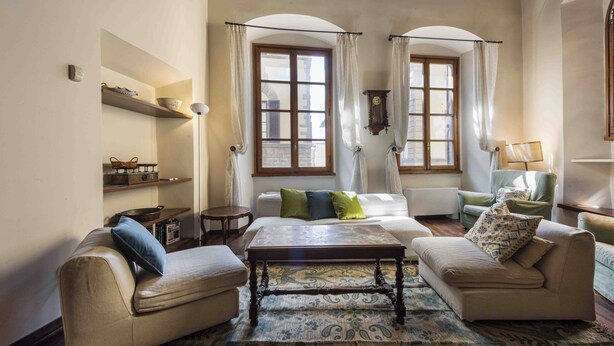 Ein Wohnzimmer mit zwei Sofas, einem Couchtisch und einem gemusterten Teppich. Zwei Fenster mit Vorhängen, eine Regal mit Dekoration und eine Lampe auf einem Beistelltisch.