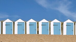 Schiera di sei cabine sulla spiaggia, con porte blu, pareti bianche e terreno sabbioso davanti.