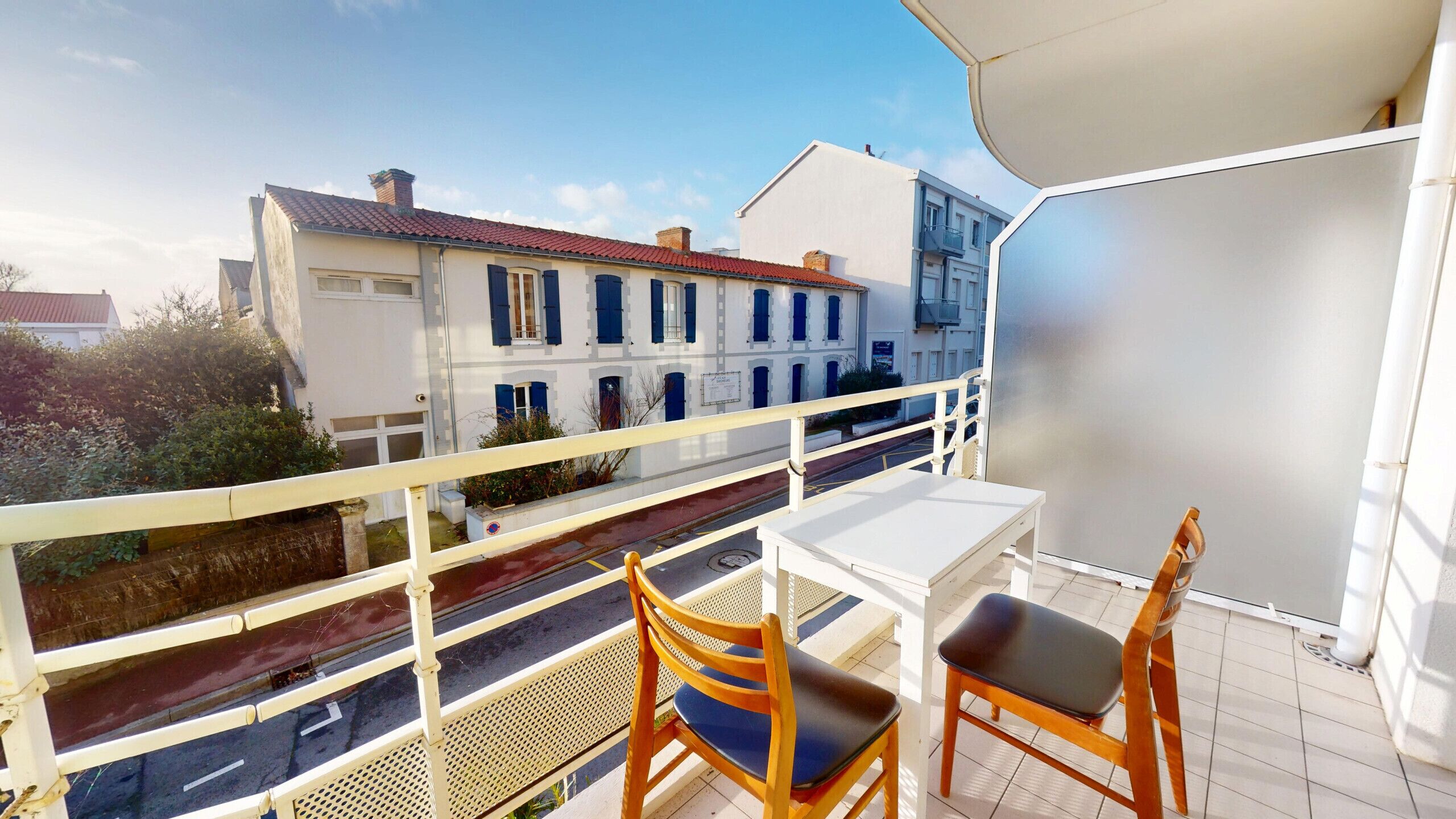 A balcony with a white table and two wooden chairs, overlooking a street with buildings and a clear sky.
