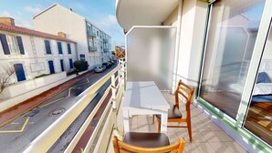 Un balcone con un tavolo bianco e due sedie di legno, affacciato su una strada con auto parcheggiate e palazzi.