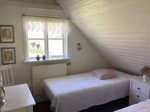 A bedroom with a sloped ceiling, a window with lace curtains, a radiator, a bedside table with a lamp, and a bed with a striped pillow. - 5 Star Holiday Home in Ansager (Ansager)