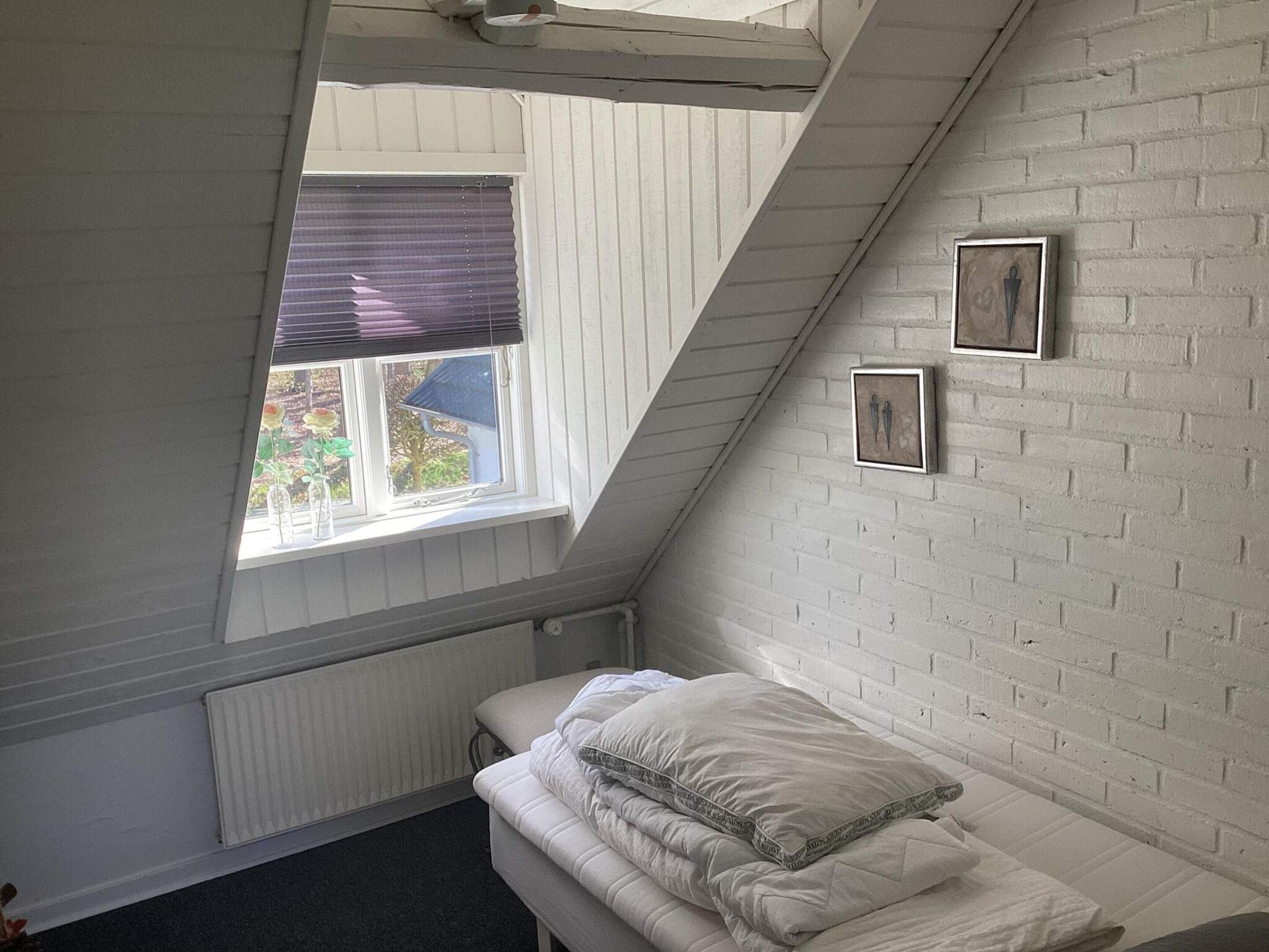 Een slaapkamer met een schuine plafond, een raam met jaloezieën, een radiator, een bed met kussens en twee ingelijste foto’s aan de muur.