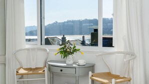 Une pièce avec deux chaises blanches, une petite table sur laquelle se trouve un vase de fleurs, une tasse et un verre de jus d’orange. Il y a des serviettes et un panier sur le sol.