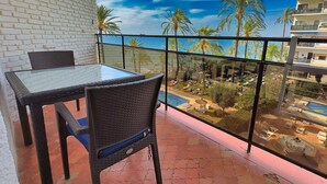 Un balcon avec une table et des chaises en verre, donnant sur un espace piscine agrémenté de palmiers et sur un bâtiment en arrière-plan.
