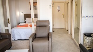 Une chambre d’hôtel avec un lit, un fauteuil marron, un canapé et une poubelle.