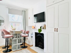 A modern kitchen with white cabinetry, a flat-screen TV, a fireplace, and pink bar stools. - The Hammersmith River studio- central, cosy and well designed (London)