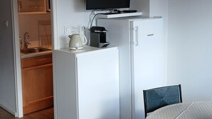 Ett litet rum med ett vitt kylskåp, en liten kaffebryggare, en tv som hänger ovanför, ett matbord med en duk med prickmönster och en stol.
