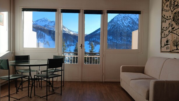 Ett rum med ett glasbord och stolar, en beige soffa och utsikt över snötäckta berg genom stora fönster.