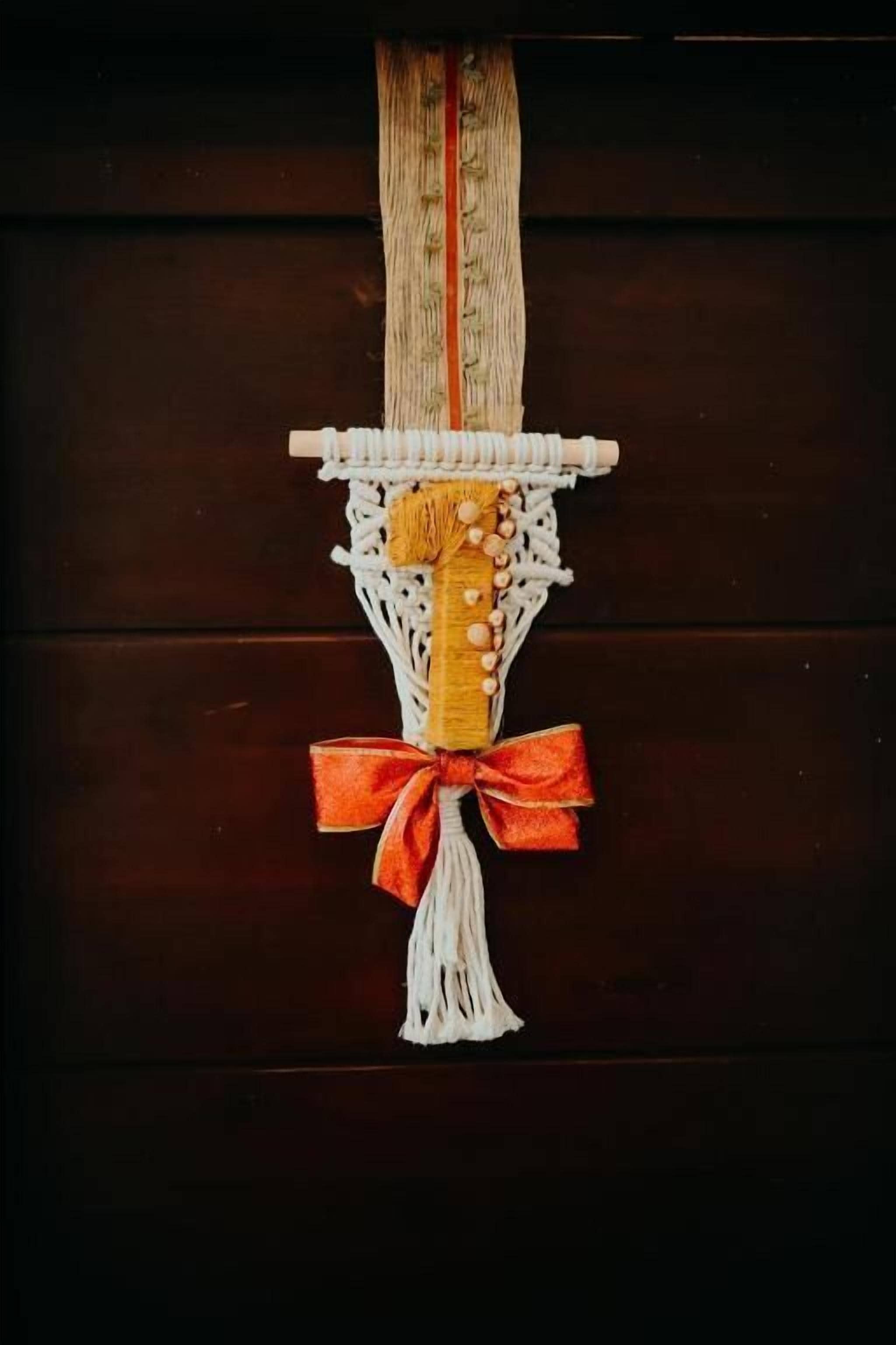 A macrame wall hanging with a central wooden block, white macrame, and an orange ribbon.