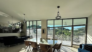 A spacious living area with a dining table, chairs, a sofa, and large windows offering a view of greenery and a distant landscape. - Spacious 6 & 8 bedroom Chalet Vista al Mar with AC and solar power in Aguadilla (aguadilla)