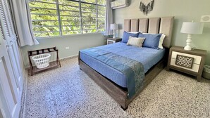A bedroom with a large window, a bed with blue bedding, two bedside tables with lamps, and a wall-mounted air conditioner. - Spacious 6 & 8 bedroom Chalet Vista al Mar with AC and solar power in Aguadilla (aguadilla)