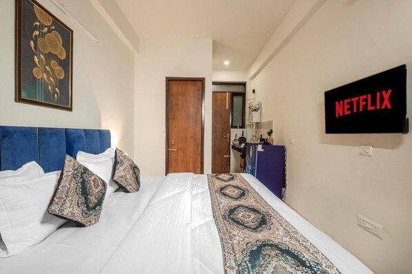 A hotel room with a bed, a blue headboard, a patterned blanket, a wall-mounted TV displaying 'NETFLIX', and a door leading to a bathroom. - COZY COVE (Dera Bassi)
