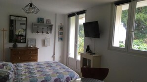 A bedroom with a bed, a round table, a chair, a TV, and a window with a view of greenery. - Les Studios de la Côte Roz: Voyageur Solo, (Perros-Guirec)