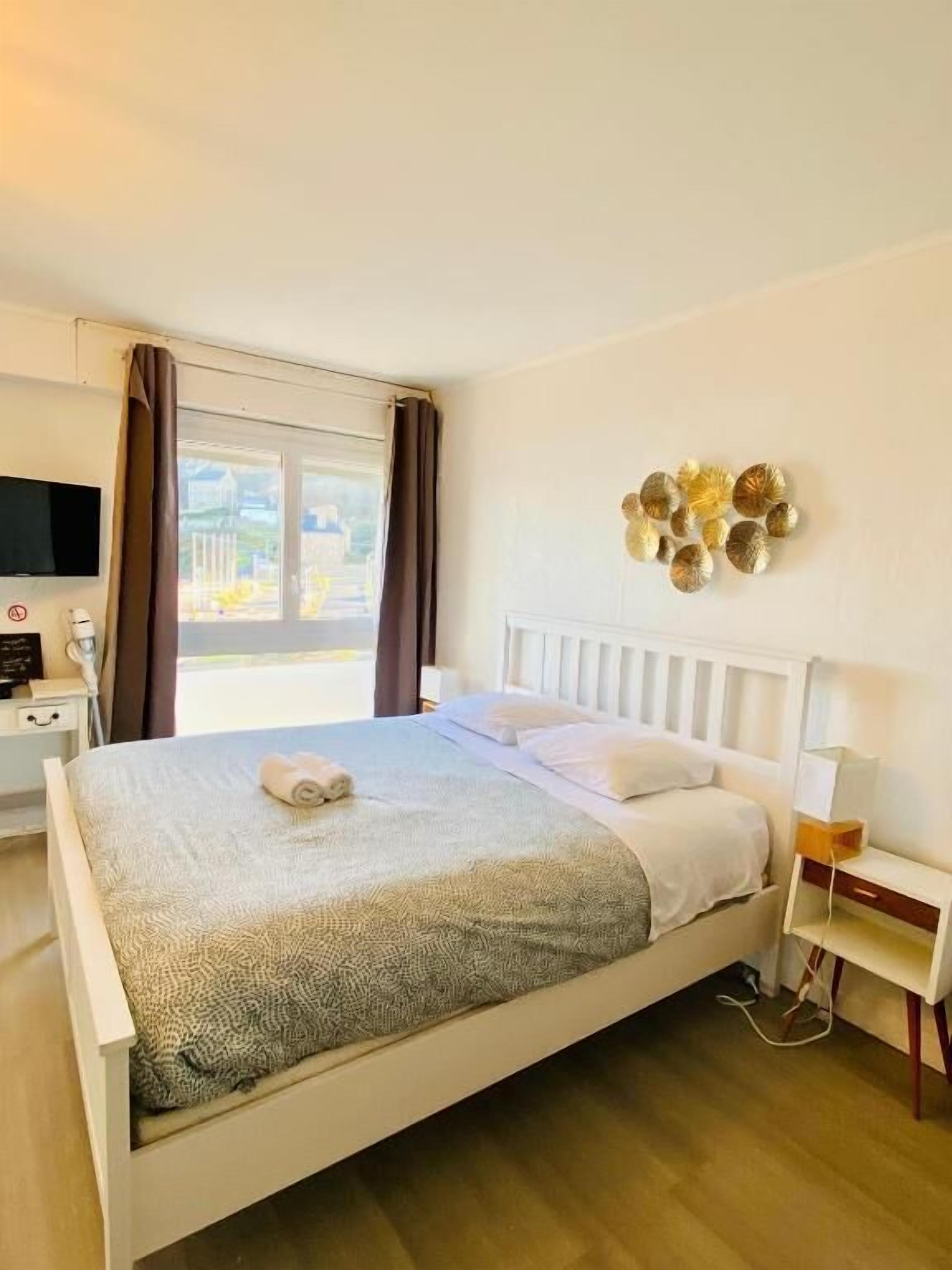 Ein ordentlich eingerichtetes Hotelzimmer mit Bett, Nachttisch, Fernseher und einem Fenster mit Vorhängen.