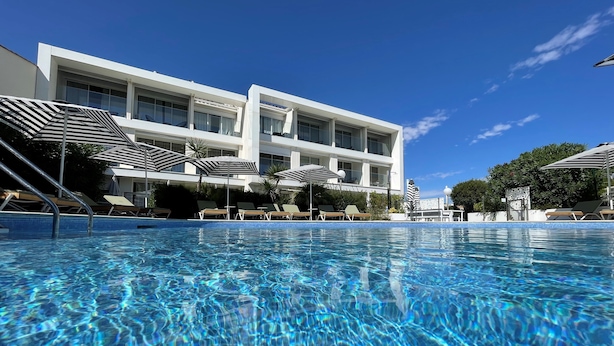 Une piscine aux eaux cristallines, dotée de chaises longues et de parasols, un bâtiment moderne aux grandes fenêtres et un ciel bleu azur.