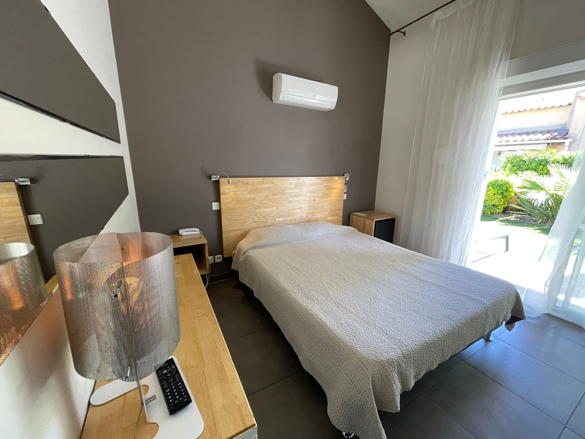 Une chambre d’hôtel moderne avec une tête de lit en bois, un grand lit recouvert d’une couverture grise, une table de chevet avec une lampe et une télécommande, et une vue sur la verdure à l’extérieur.