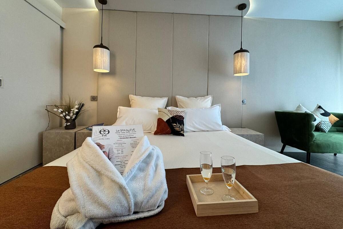 Habitación de hotel moderna con una cama grande, dos lámparas colgantes, un sillón verde y una bandeja con vasos.