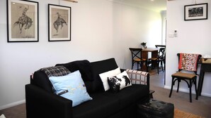 A living room with a black sofa, a coffee table, and a dining area with chairs and a table. There are framed pictures on the wall. - THE QUARTERS #1 Centrally located modern 2 bed unit featuring stylish decor (East Tamworth)