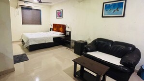 A hotel room with a bed, a black leather sofa, a small table, a ceiling fan, and a window with blinds. - Hotel Silver Leaf (Panchgani)