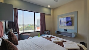 Una camera d'albergo con un letto, una TV a parete, una scrivania con un bollitore e vista sulla città attraverso ampie finestre.