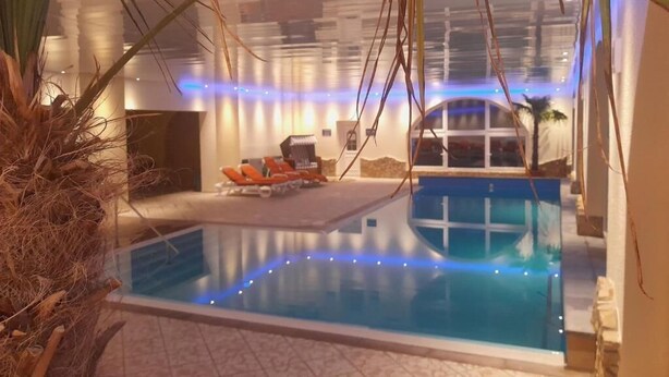 An indoor swimming pool with a glass barrier, surrounded by lounge chairs and a palm tree. - Carea Harz Hotel Allrode (Thale)