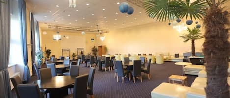 A spacious hotel lobby with multiple dining tables and chairs, a seating area with couches, and a large palm tree.