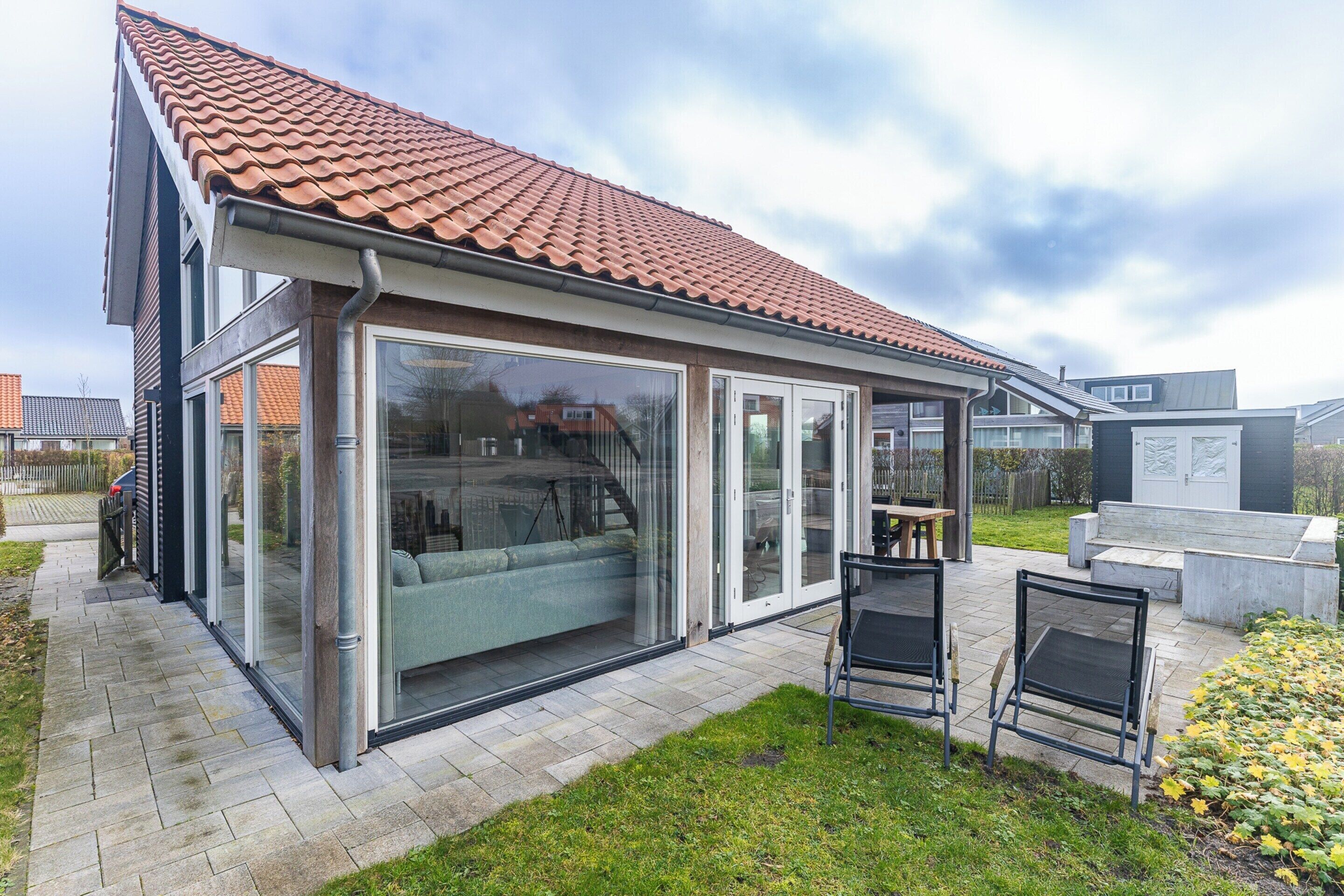 Une maison moderne avec un toit en tuiles, de grandes portes vitrées et un patio aménagé avec du mobilier d’extérieur.