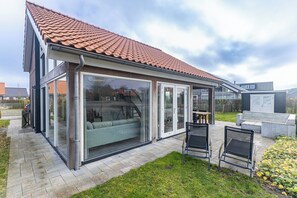 Une maison moderne avec un toit en tuiles, de grandes portes vitrées et un patio aménagé avec du mobilier d’extérieur.